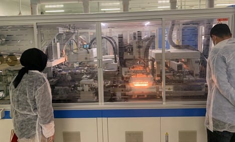 Two inspectors in cleanroom suits observing a robotic production line through a glass partition
